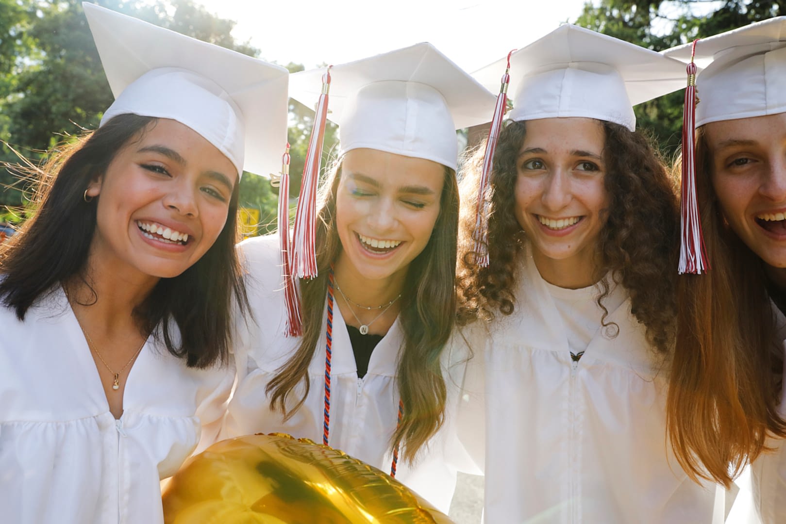 Group portrait of 4 teenage girls on their graduation day. They are all wearing white caps and gowns. They are standing outside on a street in their neighborhood. They are all smiling and having a wonderful time. There is a beautiful backlight on them. Waist up portrait.