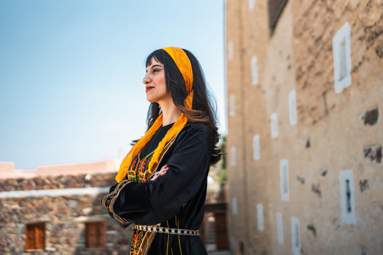 A young Middle Eastern woman in traditional Saudi dress is standing confidently at Al-Abu Sarrah Palace. She is smiling and looking into the distance, surrounded by historic stone architecture and bright daylight.