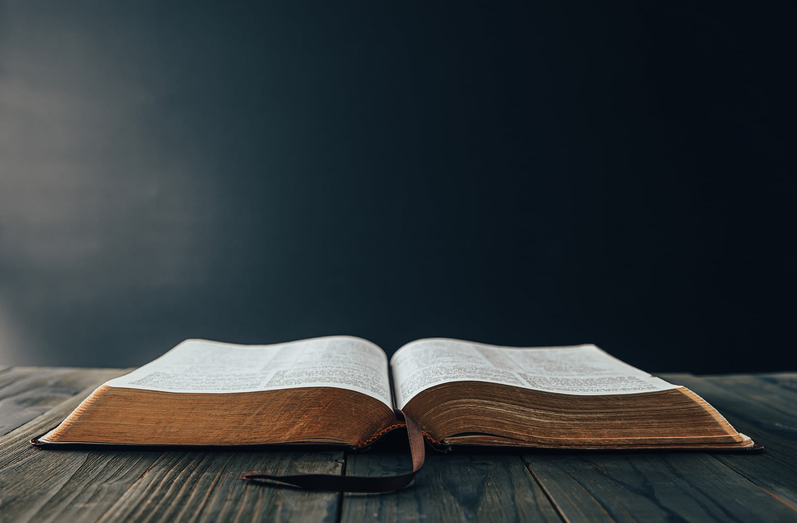 Open Bible on a wooden table against a dark background with a little light coming from the left frame.