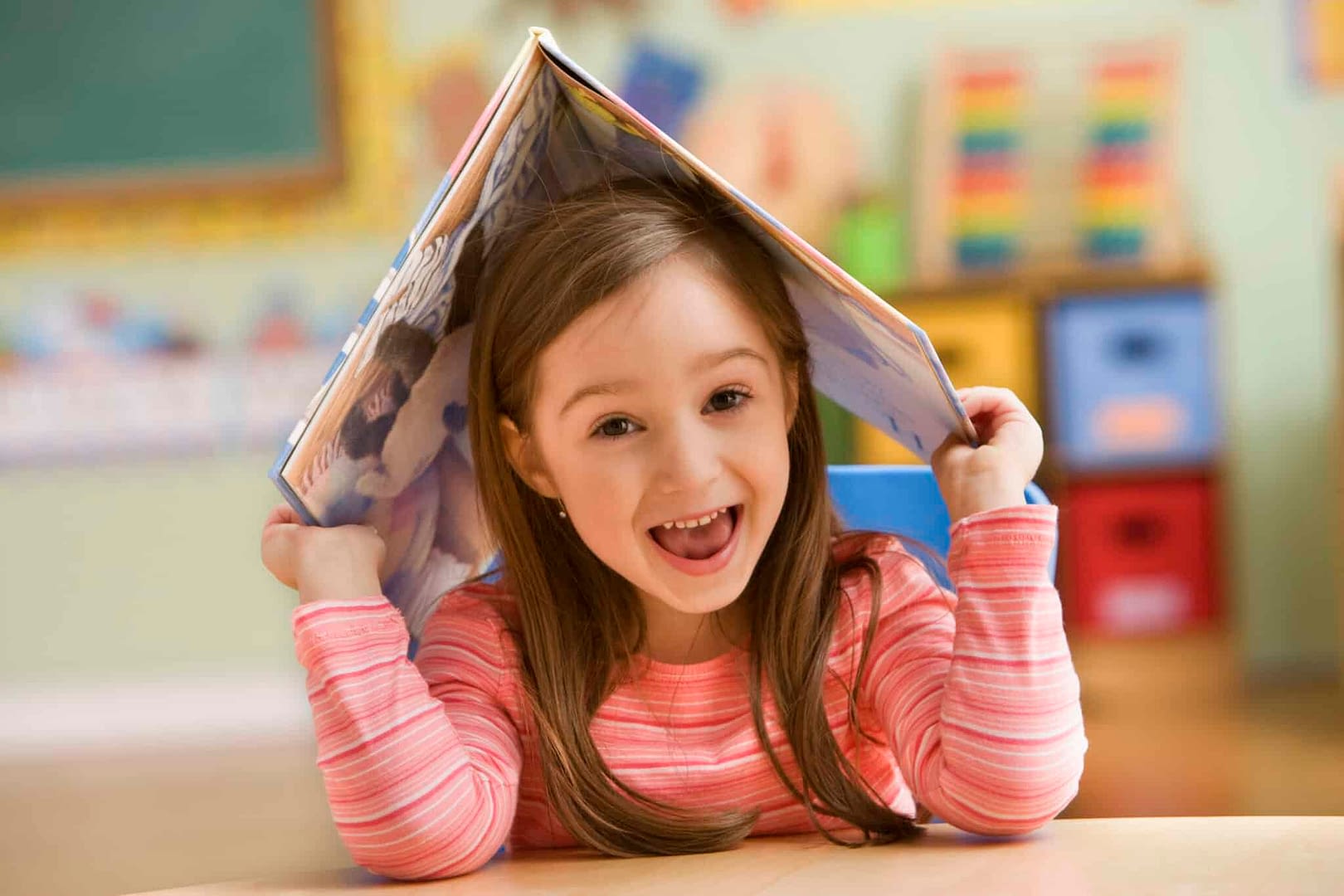 Girl (4-5) holding open book on head and smiling with open mouth